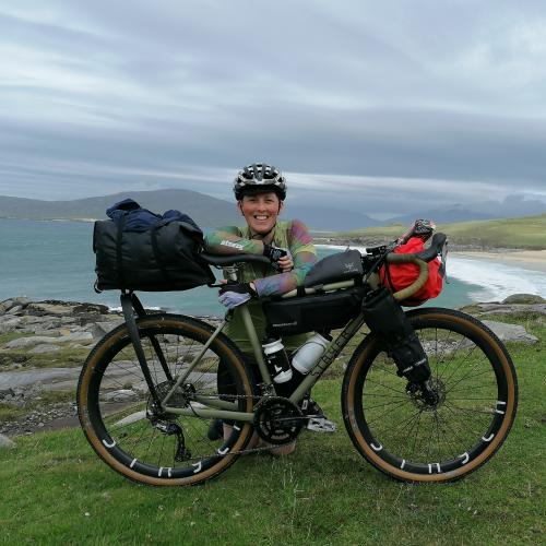 Iona kneels behind her green bike which is set up with bikepacking bags. She wears a helmet, green jersey, gloves and black shorts. A beach is in the background.