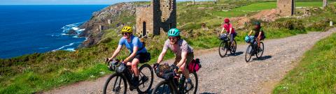 Cyclists riding along a coastal gravel trail.  There are ruins and the sea in the background.
