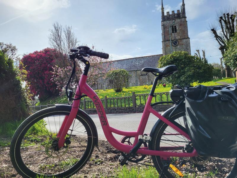 A pink Wisper e-bike with packed pannier is in the foreground with a country church in the background
