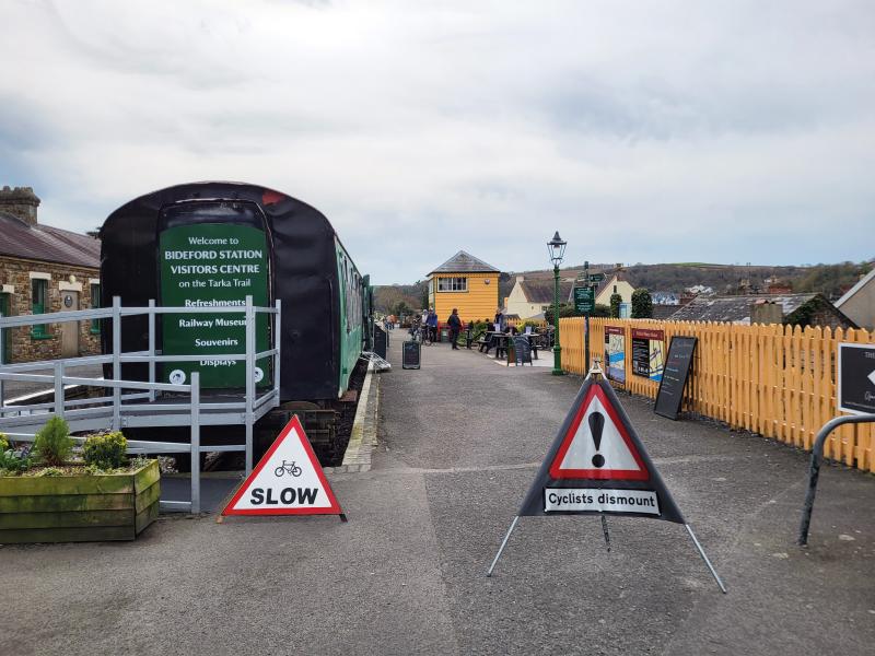 A disused railway station with an old carriage car that’s been turned into a visitor centre and café. Station buildings and people walking, cycling and sitting at picnic tables can be seen in the background. In the foreground are 'Cyclists slow' and 'Cyclists dismount' signs