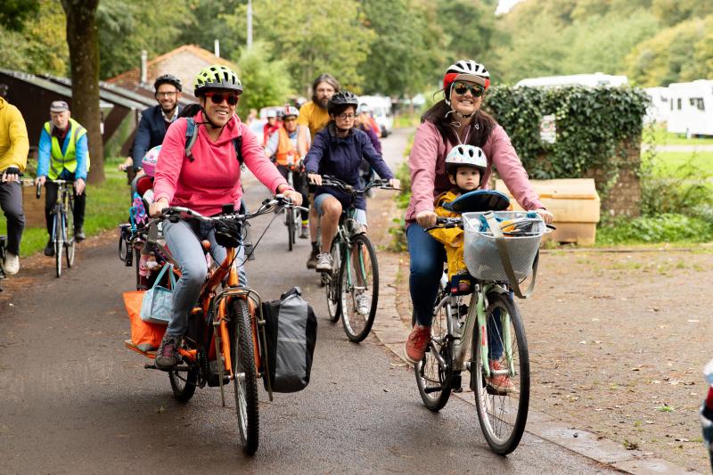 A group of people are cycling on a paved path. Two women are at the front. One is on an orange cargo bike with loaded panniers, the other a hybrid with a basket on the front and a child on a seat on the top bar.