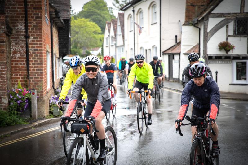 A group of people is cycling in the rain through a village. They're wearing cycling shorts and waterproofs.