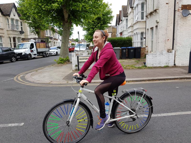 A person is riding a white hybrid bike. They are wearing leggings and a sweatshirt and trainers with rainbow laces. Their bike has rainbow spokey dokies on the spokes