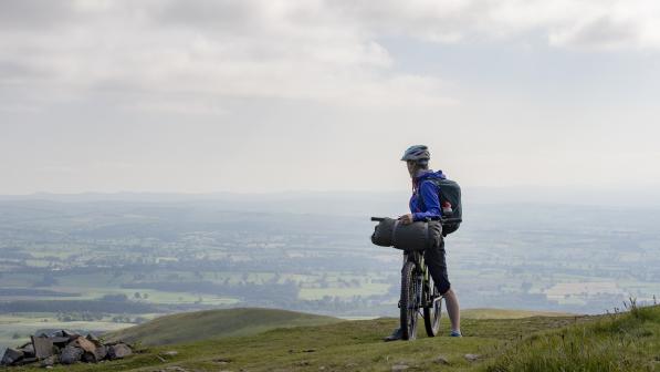 A cyclist straddling her bike. She is looking back at rolling hills behind her.