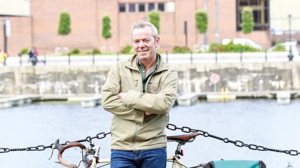 A man is leaning against a touring bike in front of a chain link fence on a path next to a river. He's wearing jeans and a jacket