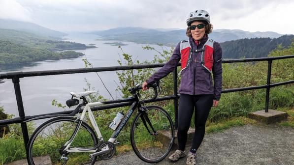 A woman is standing on a bridge next to a metal fence over a loch. She is holding a black and white road bike and is wearing warm cycling kit and a helmet