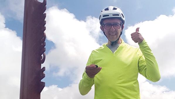 A man in a bright yellow cycling jersey and cycling helmet is standing next to a sign and giving a thumbs up