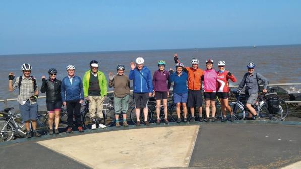 A group of 12 cyclists (men and women) are standing on a seaside promenade with their bikes. The sea is behind them. They're wearing a mix of cycling kit and normal clothes. A few are waving at the camera