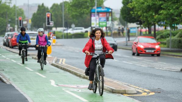 A woman in a red coat with long hair is cycling on a segregated cycle path next to a road. It's been raining and everything is wet. There are two more people on bikes behind her