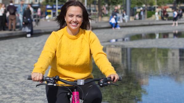A woman wearing a bright yellow jumper smiles as she cycles along a path beside a reflective water feature in a city square, with people and buildings in the background.