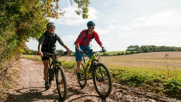 Two people are riding mountain bikes on a muddy trail in the countryside. They are wearing shorts and T-shirts and cycle helmets
