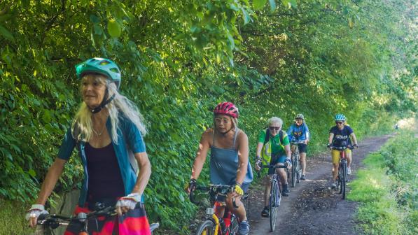 A group of women is cycling along an off-road track in the country. It's muddy. They're on mountain bikes and wearing cycling kit