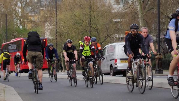 A big mixed group of people are cycling on a dedicated cycle path next to a busy London road