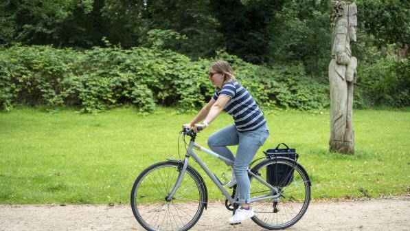 A woman in jeans and a stripy T-shirt is riding a flat-bar hybrid bike with a pannier on it on a gravel path in a park