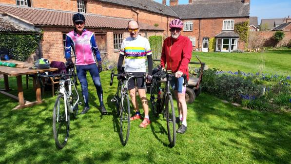 Three men in cycling kit are standing holding their road bikes in a back garden