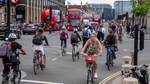 Lots of cyclists are riding on a busy London road with buses in the background. They're on all different kinds of bikes including hire bikes and folding bikes. They're wearing normal clothes.