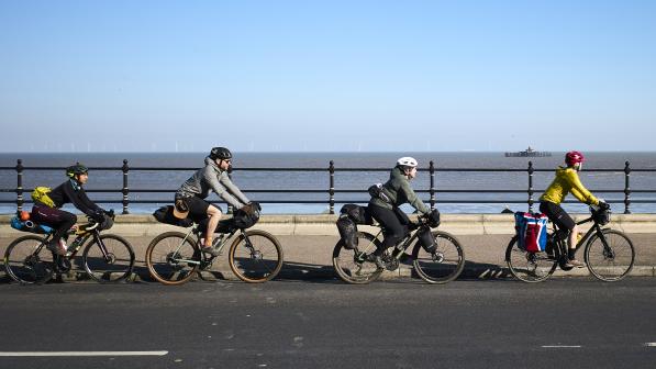 Four people are cycling in a line along a road with the sea in the background. They are all wearing cycling clothing and helmets. The sun is shining. Their bikes all have loaded panniers and bags.