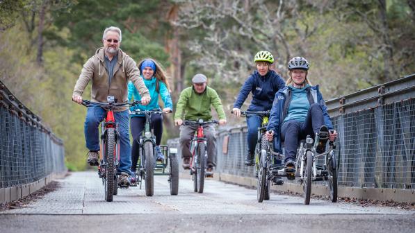 A group of people on a variety of cycles including trikes are cycling across a bridge
