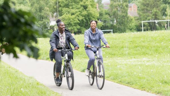 Two women in normal clothes are riding along a paved path in a park on flat bar bikes