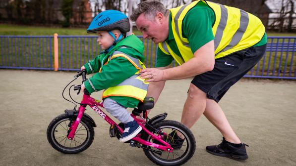A child is riding a children's bike with pedals with an adult instructor standing behind with a supportive hand on the child's back