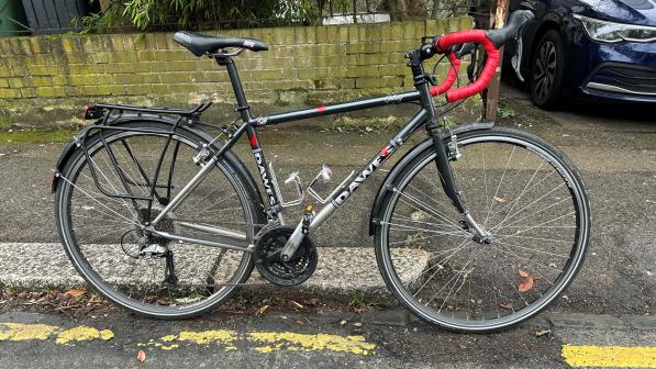 A black Dawes Galaxy touring bike with red bar tape is propped against a curb with the front wheel in a pothole