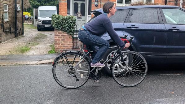 A woman is cycling a touring bike along a wet urban road. She's wearing a pair of jeans and a sweater