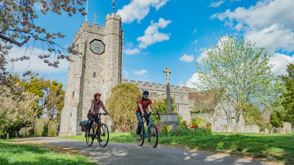 Two people on touring bikes with luggage are cycling past a country church in spring