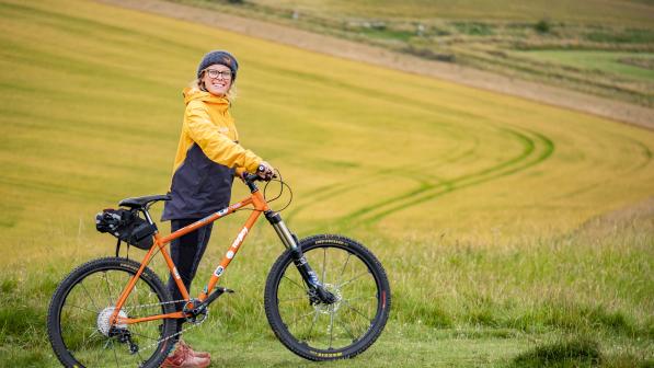 Alex Cuppleditch standing in an open field with a smile, wearing an orange and black jacket and holding an orange mountain bike. Rolling hills and a winding path can be seen in the background, under a cloudy sky.
